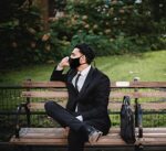 Man in a suit wearing a black cloth mask on a park bench