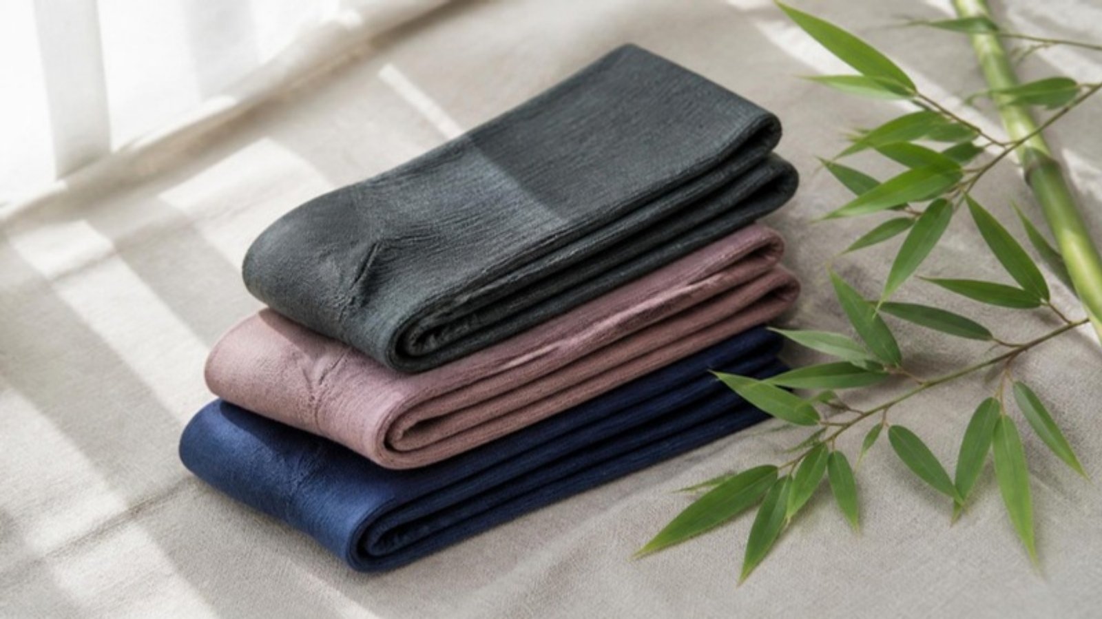 Three folded knee-high bamboo compression socks in charcoal gray, dusty rose and navy on a linen surface beside green bamboo leaves and a bamboo stalk