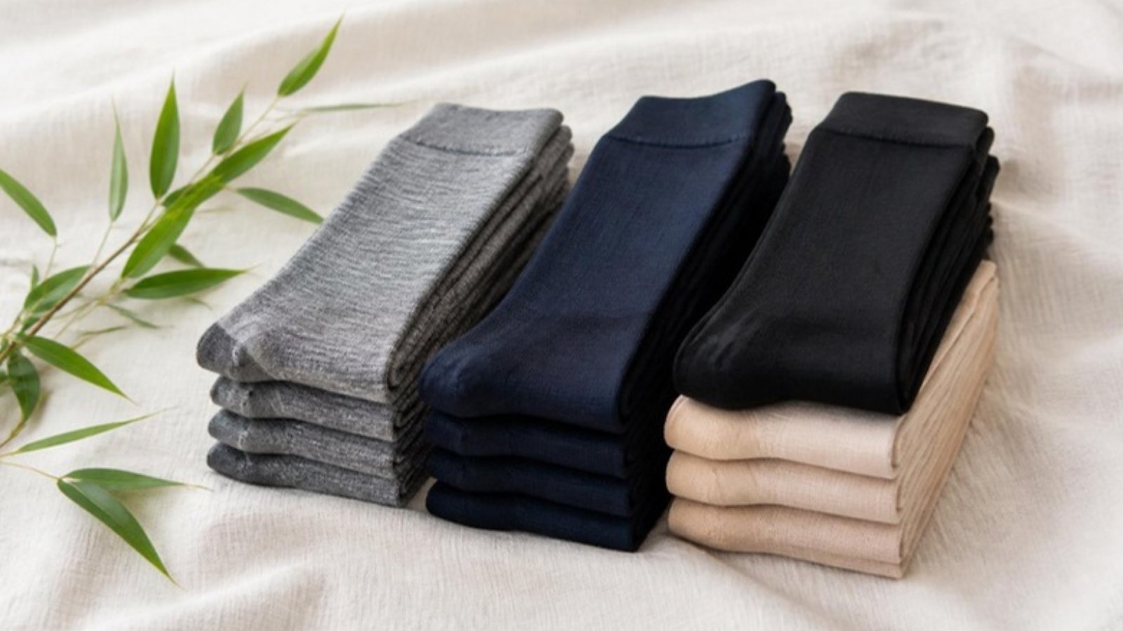 Stacks of folded knee-high socks in gray, navy, black and beige on a linen cloth beside green bamboo leaves