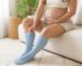 Pregnant woman sitting on a light sofa pulling on light blue knee-high compression socks