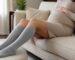 Pregnant woman in a beige knit dress resting on a sofa with her legs on an ottoman, wearing light gray knee-high compression socks