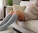 Pregnant woman in a beige knit dress resting on a sofa with her legs on an ottoman, wearing light gray knee-high compression socks