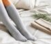 Legs in soft light gray knee-high socks resting on a white bed beside a folded beige pair and a bamboo sprig