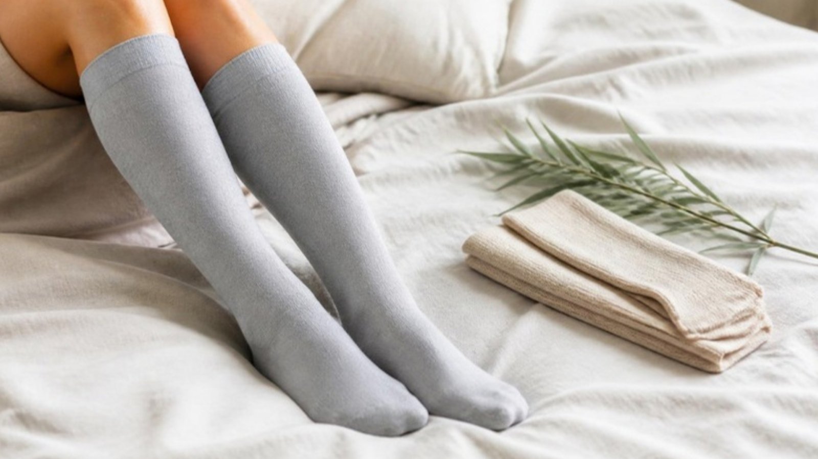 Legs in soft light gray knee-high socks resting on a white bed beside a folded beige pair and a bamboo sprig